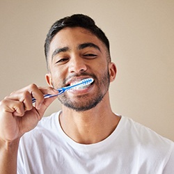 Man in white shirt brushing his teeth with a tan background